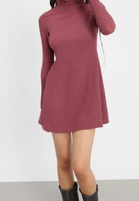 Långärmad, vinröd klänning med strukturväv, hög halsringning och volangdesign, som slutar ovanför knäet, tillsammans med knähöga svarta stövlar.