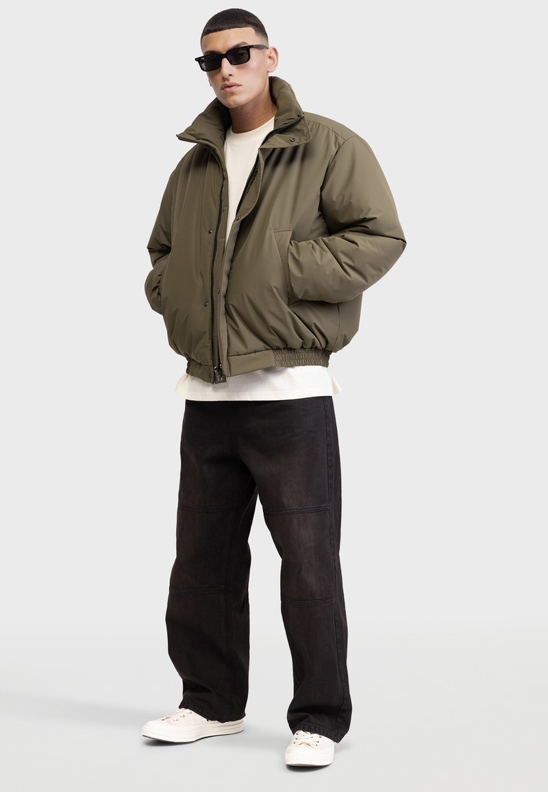 Giacca imbottita verde oliva con colletto alto, zip frontale e chiusura a scatto. Indossata sopra una maglietta bianca e abbinata a pantaloni in denim scuro e sneaker bianche.