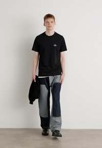 Čierne bavlnené tričko s malým bielym potlačou, skombinované s patchwork džínsami v rôznych odtieňoch denimu. Model drží čiernu bundu a má na sebe čierne topánky.