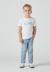 T-shirt blanche ornée d'un logo Levi's bleu, associée à un jean bleu clair et des baskets blanches avec des accents noirs. Design décontracté.