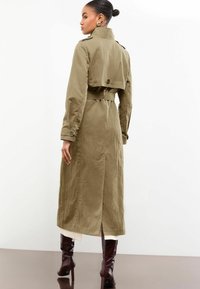 Vrouw met een knot in haar haar, gekleed in een met een riem afgewerkte olijfgroene trenchcoat en bruine hakschoenen, staand en naar de zijkant kijkend tegen een witte achtergrond.