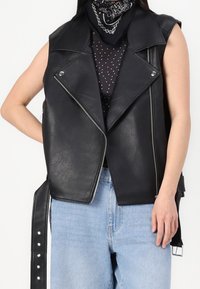 Černá vesta bez rukávů z umělé kůže s klopami, stříbrnými zipy a bočními sponami. Nositelná přes top s puntíky a světle modré džíny.