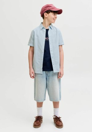 Poiss punases mütsis, helerohelise lühikese varrukaga särgiga üle mereväe graafilise t-särgi, põlveni ulatuvate teksapükste, valgete sokkide ja pruunide kingadega, naeratades külgsuunas.