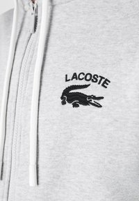 Sweat à capuche zippé gris en tissu doux, avec un logo Lacoste brodé noir représentant un crocodile. Cordons blancs au niveau du col.