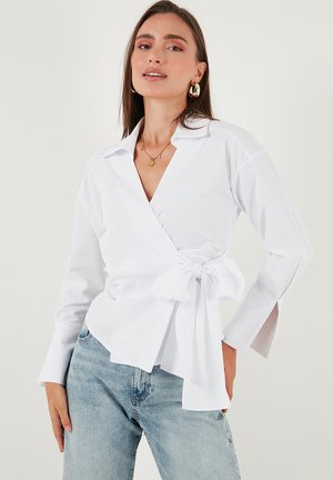 Woman wearing a white wrap blouse with a large bow on the side and light blue jeans, standing against a plain background.