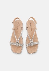 Beige mokasiinit nahkaa, joissa on neliön muotoinen varvas, hopeiset strassihihnat ja koristeellinen solmu. Säädettävä buckles-yksityiskohta nilkassa.