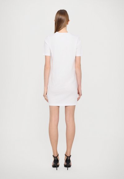Robe T-shirt blanche courte avec une coupe droite, des manches courtes et un col rond, associée à des chaussures à talons hauts noires. Texture lisse.