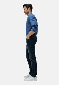 Hombre de perfil con las manos en los bolsillos, vistiendo una camisa de botones azul, jeans oscuros y zapatillas blancas, contra un fondo sencillo.