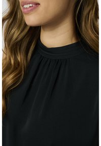 Femme aux longs cheveux bruns ondulés portant un haut noir à col montant, souriant légèrement avec les dents du haut visibles.
