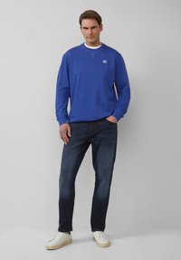 Chemise bleu à manches longues avec un col blanc, portée avec un jean foncé et des baskets blanches. La chemise arbore un petit logo sur la poitrine.
