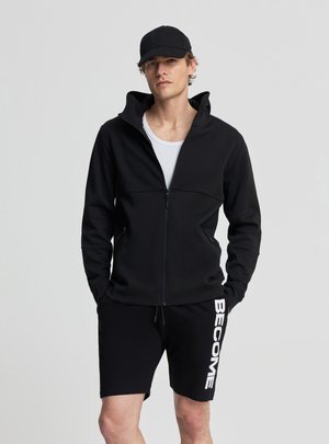 Uomo con giacca nera con zip, berretto nero, camicia bianca e pantaloncini neri con la scritta "BECOME" sulla gamba sinistra, in piedi con le mani nelle tasche.