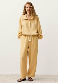 Femme aux longs cheveux bouclés portant un pull à capuche beige et un pantalon large assorti, les mains dans les poches, debout sur un sol fissuré.