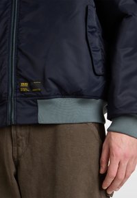Marineblaues Bomberjacke mit glatter Oberfläche, gerippten Bündchen und Saum in Hellblau, Reißverschluss und einem Vans-Label an der Seite. Braune Cordhose.