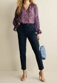 Blusa floreale trasparente viola con maniche a sbuffo, abbinata a pantaloni scuri color navy. Porta una borsa raccolta azzurro chiaro. Tacchi neutri con cinturini.