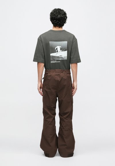 Ohlapne rjave hlače v kombinaciji s temno sivo majico z belim grafičnim tiskom na hrbtu, ki prikazuje smučarja.