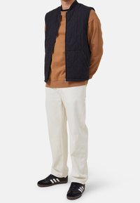 Granatowa pikowana kamizelka ze stojącym kołnierzem na brązowej bluzie, w zestawie z kremowymi spodniami i czarnymi trampkami z białymi paskami.