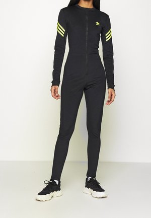 Czarny kombinezon z długim rękawem zapinany na froncie na zamek, z trzema żółtymi paskami na każdym ramieniu oraz dopasowanym krojem. Do tego czarno-białe sneakersy.