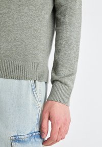 Grå, strukturerad stickad tröja med ribbade ärmslut, kombinerad med ljusblå jeans. Nærbild av arm och hand mot en enfärgad bakgrund.