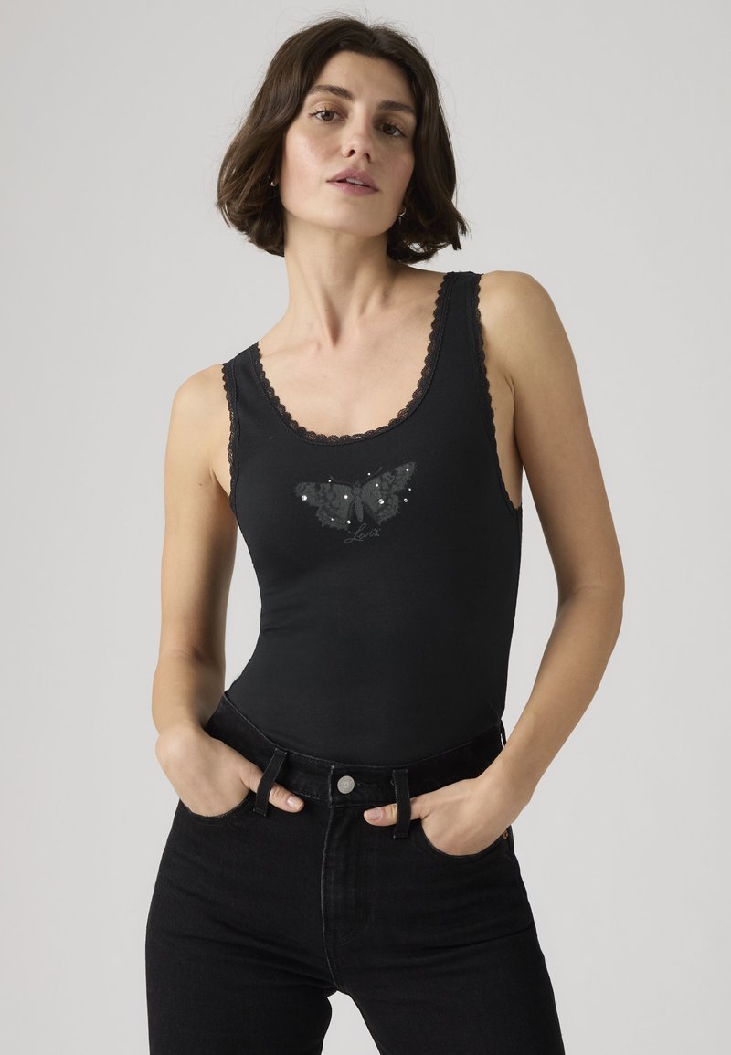 Vrouw draagt een zwart mouwloos topje met kanten rand en vlindermotief, gecombineerd met zwarte high-waisted jeans, staand met handen in de zakken.