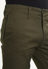 Pantaloni verde oliva realizzati in morbido tessuto di cotone, caratterizzati da un design aderente, chiusura con bottone e passanti per cintura standard.