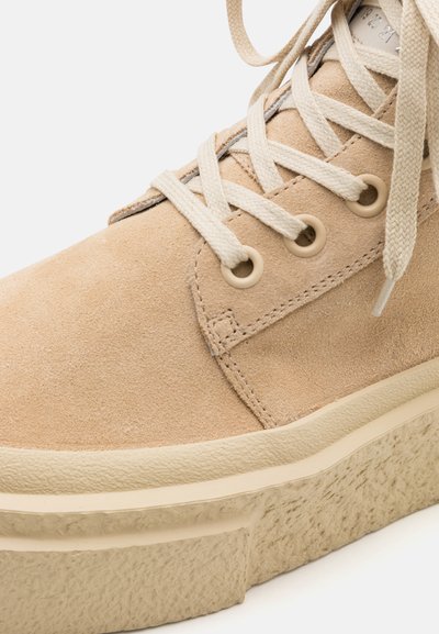 Beige suede sneaker with a textured sole, featuring round beige eyelets, flat beige laces, and a minimalist design.
