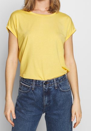 Frau trägt ein gelbes Kurzarmoberteil, in eine hoch taillierte dunkelblaue Jeans gesteckt, steht vor einem schlichten Hintergrund.