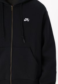 Felpa nera con zip realizzata in un tessuto morbido, dotata di tasca frontale, cappuccio con lacci e un piccolo logo Nike SB bianco sul petto.