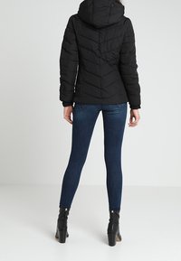 Svart kviltad puffjacka med huva, med chevronstickning och en åtsittande silhuett. Buren över mörka smala jeans och ankelstövlar.