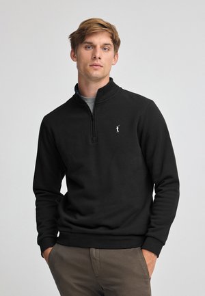 Zwarte longsleeve pullover met een kwart-rits kraag, voorzien van een klein logo op de linkerborst, gemaakt van een zachte stof met een gladde textuur.