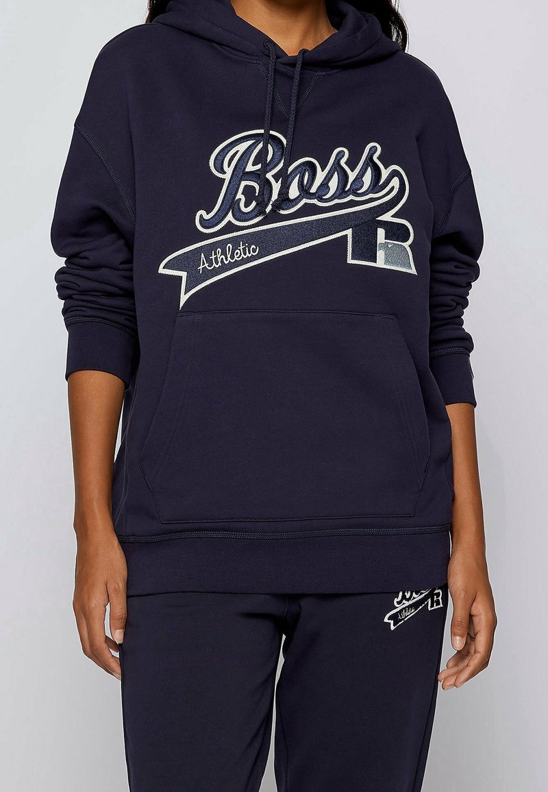 Personne portant un sweat à capuche bleu marine et un pantalon assorti avec le logo "Boss Athletic" en blanc et beige sur la poitrine et la cuisse.