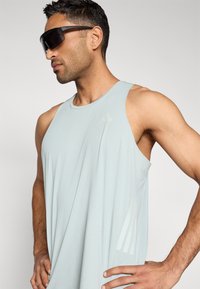 Šviesiai mėlynas be rankovių sportinis įliemenuotas marškinėlis su laisvu prigludimu ir trimis vertikaliomis juostelėmis šone. Pagamintas iš lygaus, kvėpuojančio audinio.