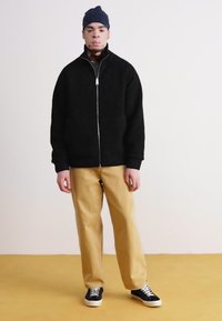 Sort strikket zip-up jakke med høj krave, parret med beige bukser og sorte sneakers, sat mod en neutral baggrund.