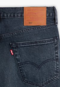 Jeans de mezclilla azul oscuro con un parche de cuero marrón que presenta texto y diseños en relieve. Etiqueta de marca roja en el bolsillo trasero. Diseño clásico.