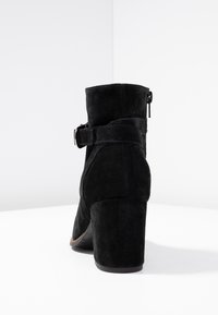 Bottes chelsea en suédine noire avec un talon épais et une fermeture éclair sur le côté, présentant une sangle décorative avec un détail de boucle. Texture lisse et bout arrondi.