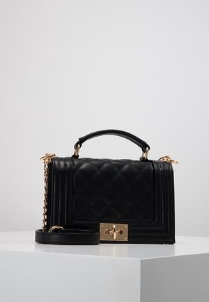 Bolso acolchado negro con asa superior, correa de cadena dorada y cierre rectangular dorado con giro sobre una superficie blanca.