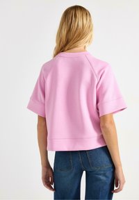 Felpa corta rosa con maniche corte e larghe e scollo rotondo. Il tessuto appare morbido e resistente. Abbinata a jeans blu a vita alta.