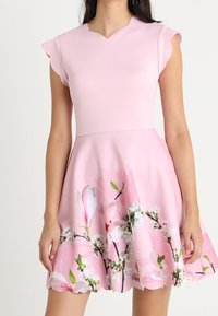 Robe A-line rose avec des manches courtes en volants, décolleté en V et imprimé floral au bas, présentant des fleurs roses et des accents verts sur un tissu lisse.
