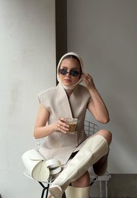 Bež brezrokavnik s strukturiranim dizajnom, bela ruto, temna sončna očala, drži pijačo, v kombinaciji z belimi škornji do kolen in torbico.