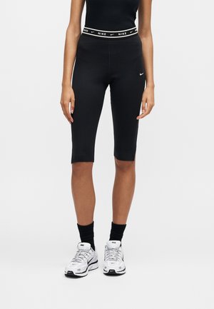 Personne portant un short de cycliste Nike noir, un haut noir, des chaussettes noires et des baskets Nike blanches sur un fond clair uni.