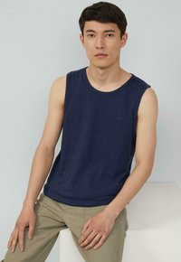 Donkerblauwe mouwloze tanktop van zachte stof, met een ronde halslijn, gecombineerd met lichtkhaki broeken, zittend tegen een neutrale achtergrond.