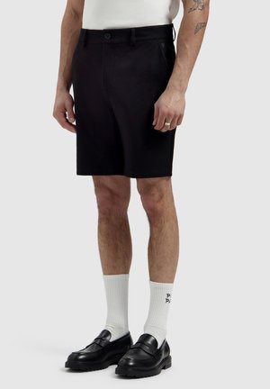 Man draagt zwarte op maat gemaakte korte broek, witte sportsokken met zwarte tekst en zwarte leren loafers, staand tegen een effen lichte achtergrond.