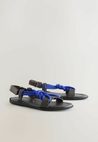 Sandales plates noires avec des brides bleues nouées et des fermetures réglables à la cheville sur un fond clair uni.