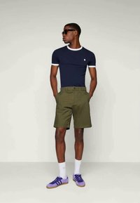 Námornícky tričko s bielym lemom, olivovo-zelené šortky a fialové tenisky. Okuliare na tvári, ruky v vreckách, stojí proti neutrálnemu pozadiu.