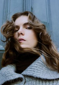 Mujer joven con cabello castaño ondulado, vistiendo un suéter grueso de punto gris y un cuello alto oscuro, sobre un fondo azul apagado.