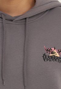 Sudadera con capucha gris con bolsillo delantero, cordón ajustable y un diseño gráfico que presenta un diseño rosa con acentos oscuros y un logo.