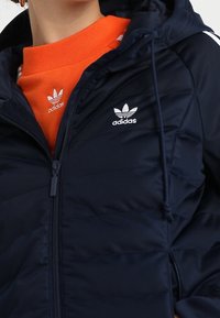 Námořnická zateplená bunda s kapucí, která má přední zip a bílé logo Adidas. Nahoře má na sobě jasně oranžovou mikinu.
