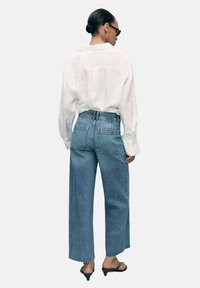 Široke traperice u izblijedjelo plavoj boji s raw rubovima, uparene s labavom bijelom lanenom košuljom. Sadrži stražnje džepove i minimalne detalje šivanja.