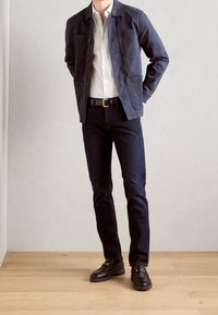 Giacca di cotone blu navy con due tasche frontali, camicia bianca, jeans di denim scuro e mocassini neri con dettagli dorati su pavimento in legno.