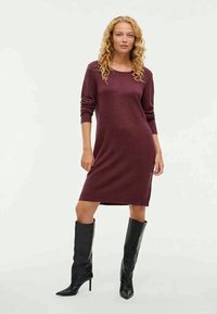 Vestido de malha bordô com mangas compridas, decote redondo e bainha canelada, combinado com botas pretas até ao joelho. Textura suave, design justo.