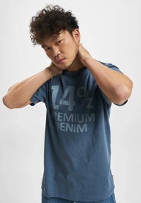 Blå bomull t-skjorte med stor lysegrå "14% PREMIUM DENIM" trykk. Korte ermer og rund hals; båret med hendene på nakken.